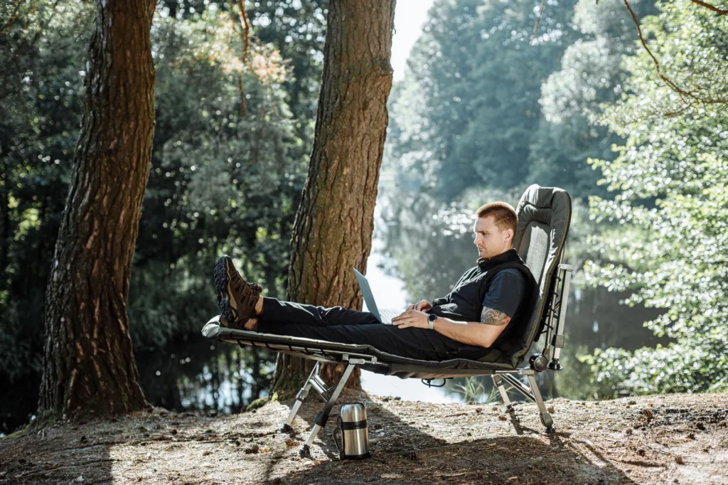 Man reclining on an extra-wide folding camping cot by a lakeside, relaxed comfort with laptop and thermos nearby